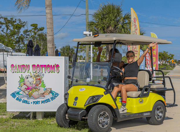 2 friends riding a Yamaha golf cart rental in Fort Myers Beach in front of Sandy Bottom's