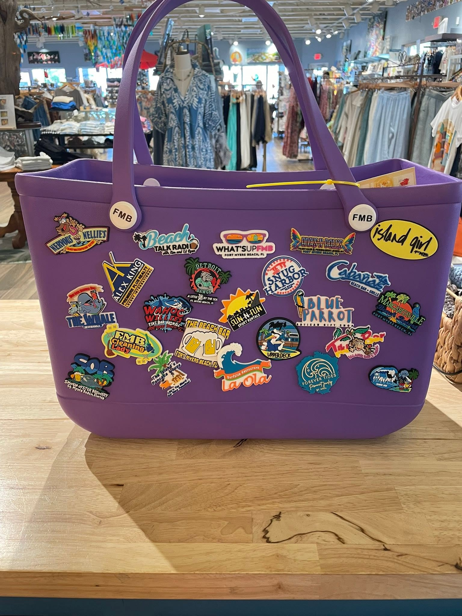 A bright purple rubber tote bag sitting on a wooden counter in a shop. The bag is covered in colorful custom charms featuring the logos of various local Fort Myers Beach businesses participating in the Island Charm Crawl.